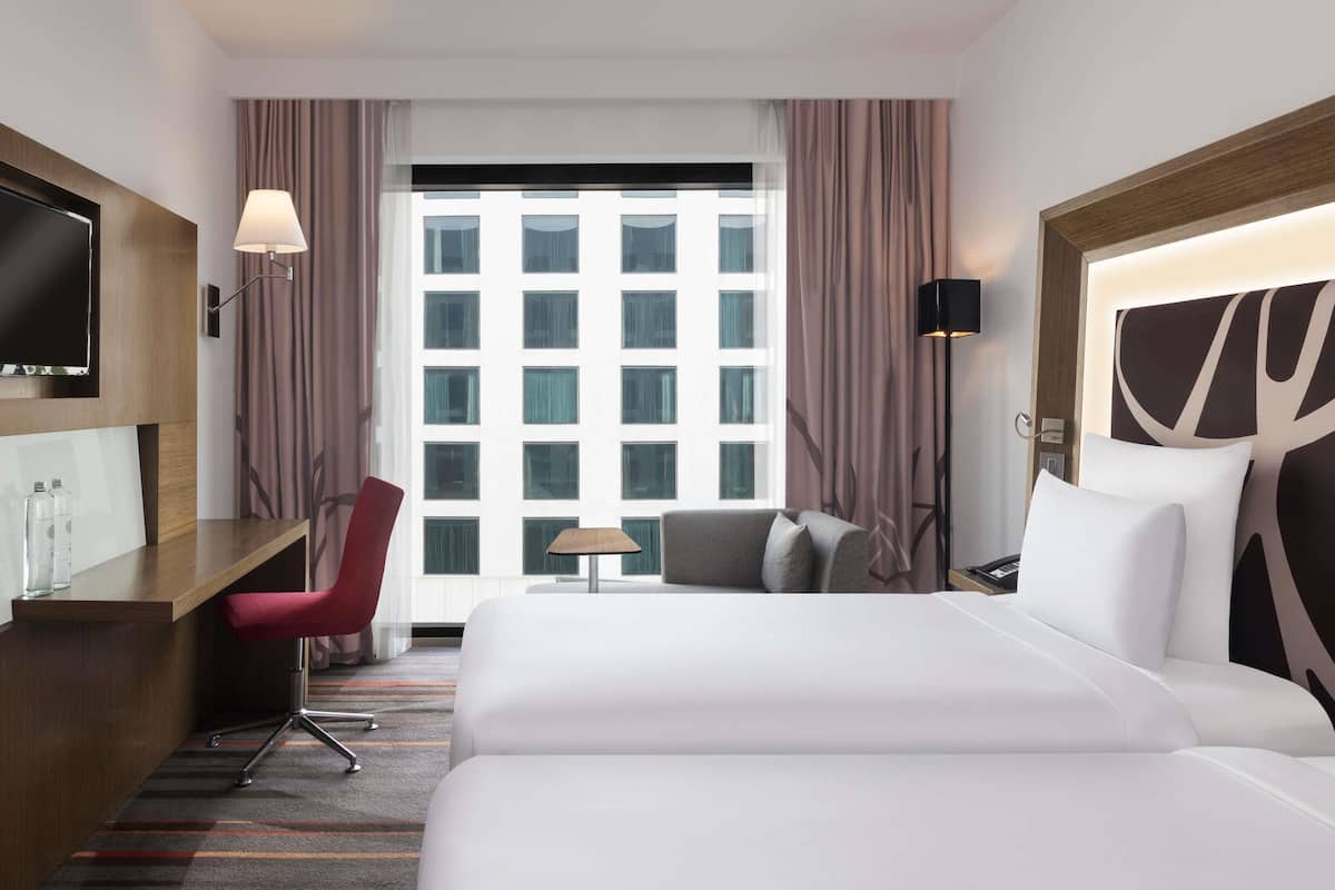 A hotel room with a bed, desk, chair, TV, lamp, and a view of a building through the window.