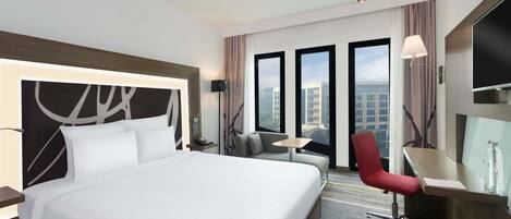 A hotel room with a large bed, a desk, a chair, a small table, and a TV.