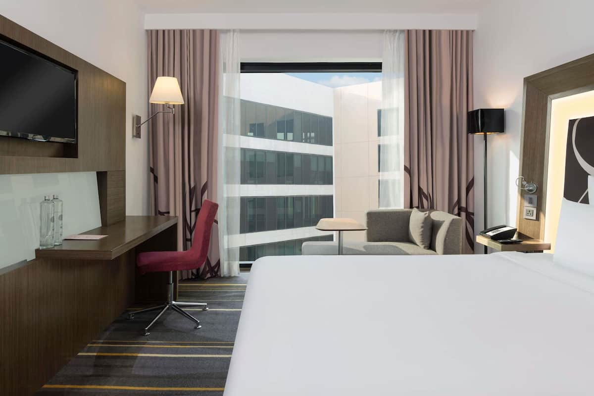A hotel room with a bed, desk, chair, TV, lamp, and large windows with curtains.