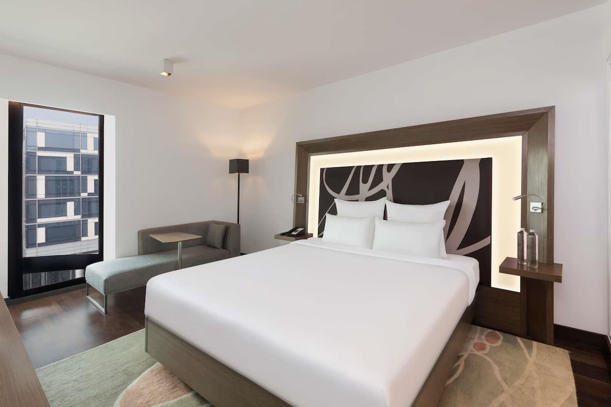 Een moderne hotelkamer met een groot bed, een slaapbank, een nachtkastje, een lamp en uitzicht op een gebouw door een raam.