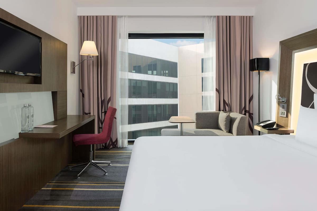 A hotel room with a bed, a desk, a chair, a TV, a lamp, and a view of a building.