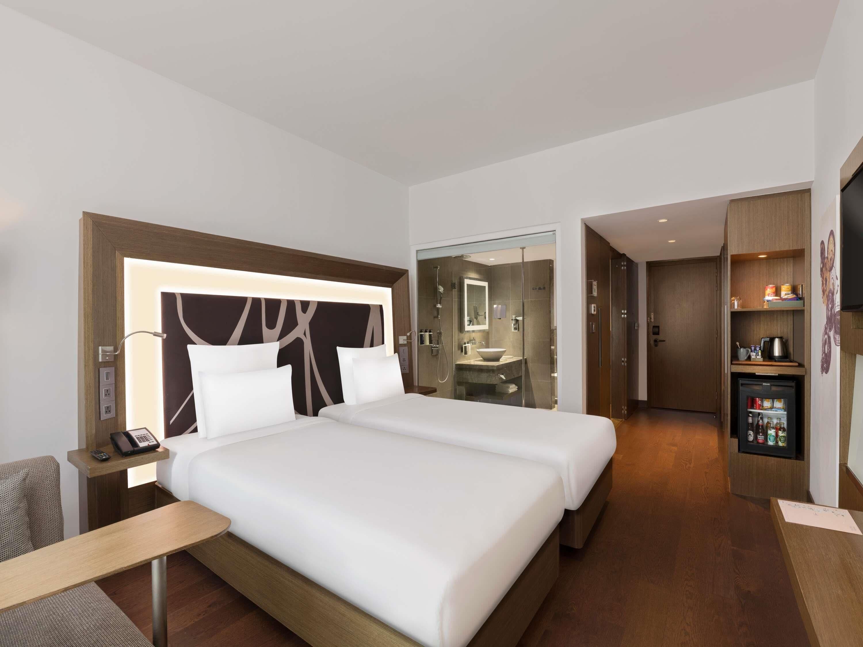 Een hotelkamer met een groot bed, nachtkastjes, een bureau, een minibar en een badkamer zichtbaar achter een glazen wand.