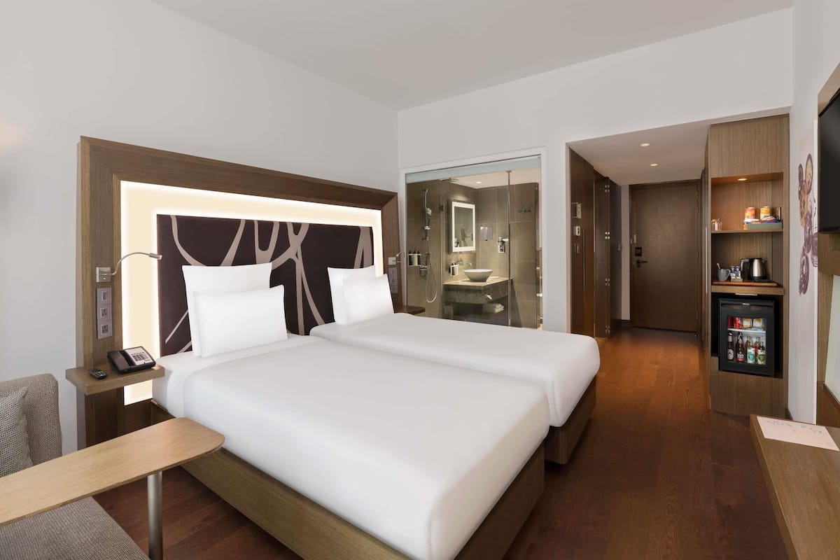 Een hotelkamer met een groot bed, nachtkastjes, een bureau, een minibar en een badkamer zichtbaar achter een glazen wand.