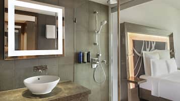 A modern bathroom with a large mirror, a vessel sink, a glass-enclosed shower, and a bed with a headboard.