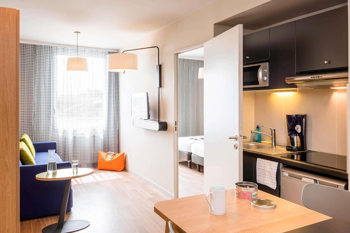 Habitació d'hotel moderna amb un sofà, una taula petita, una làmpada, un llit i una cuineta.