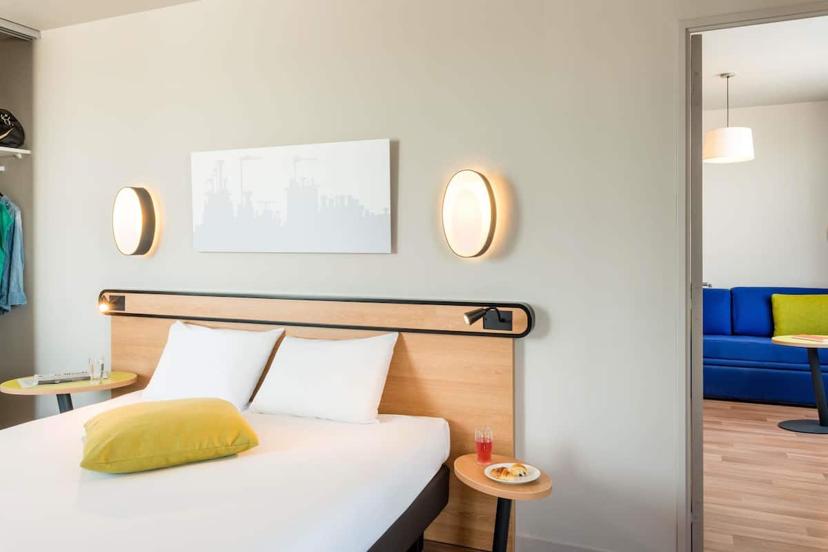Ein Hotelzimmer mit Bett, Nachttischen, Stadtbild-Kunstwerk und Wandleuchten, sowie ein Blick auf ein blaues Sofa und einen Essbereich im Hintergrund.