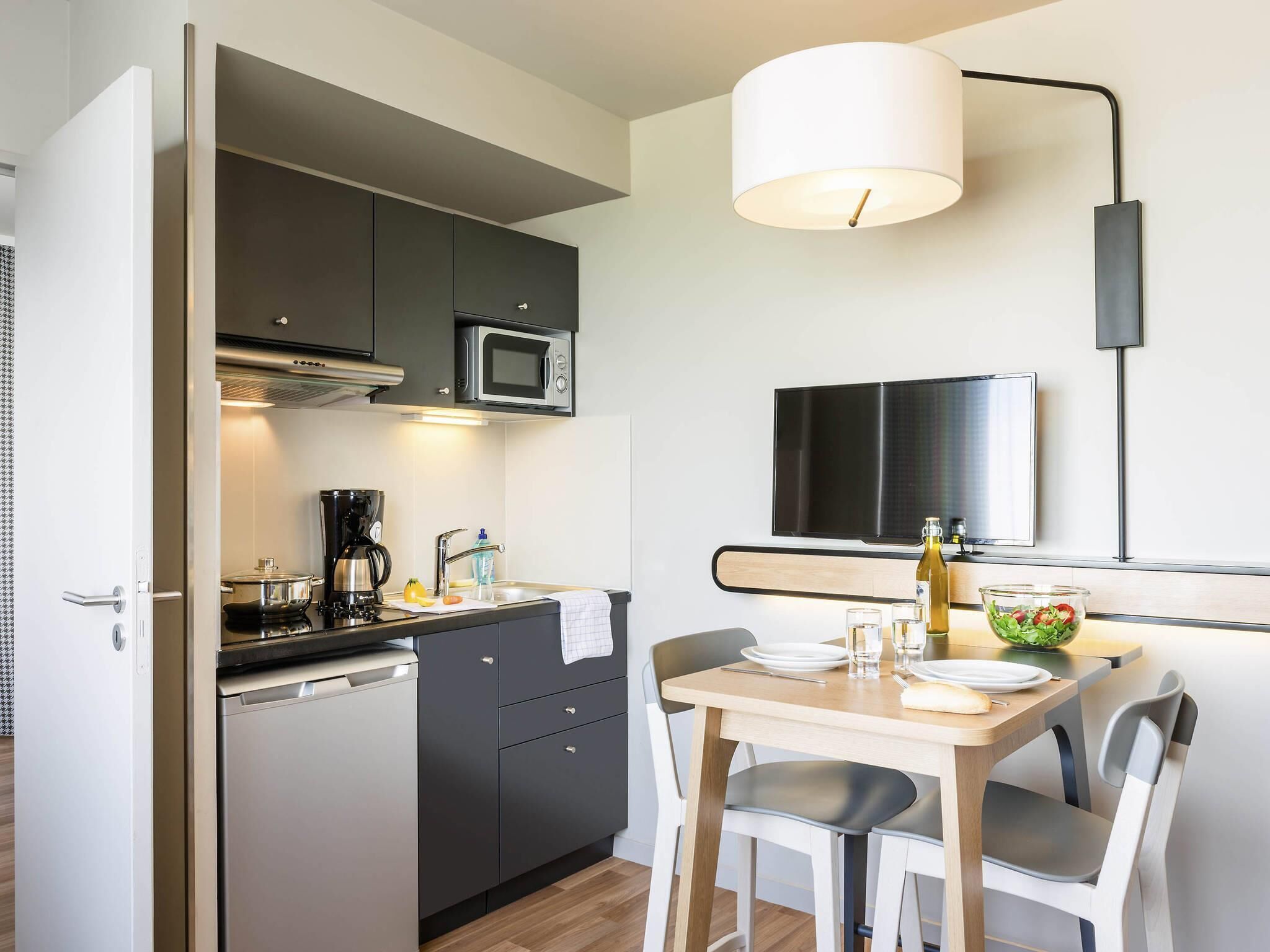 Una cocina compacta con electrodomésticos modernos, un comedor con mesa y sillas, y un televisor de pantalla plana montado en la pared.