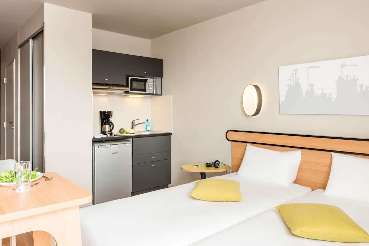 Ein Hotelzimmer mit Bett, kleinem Schreibtisch, einer Küchenzeile mit Mikrowelle und einem Stadtbild an der Wand.