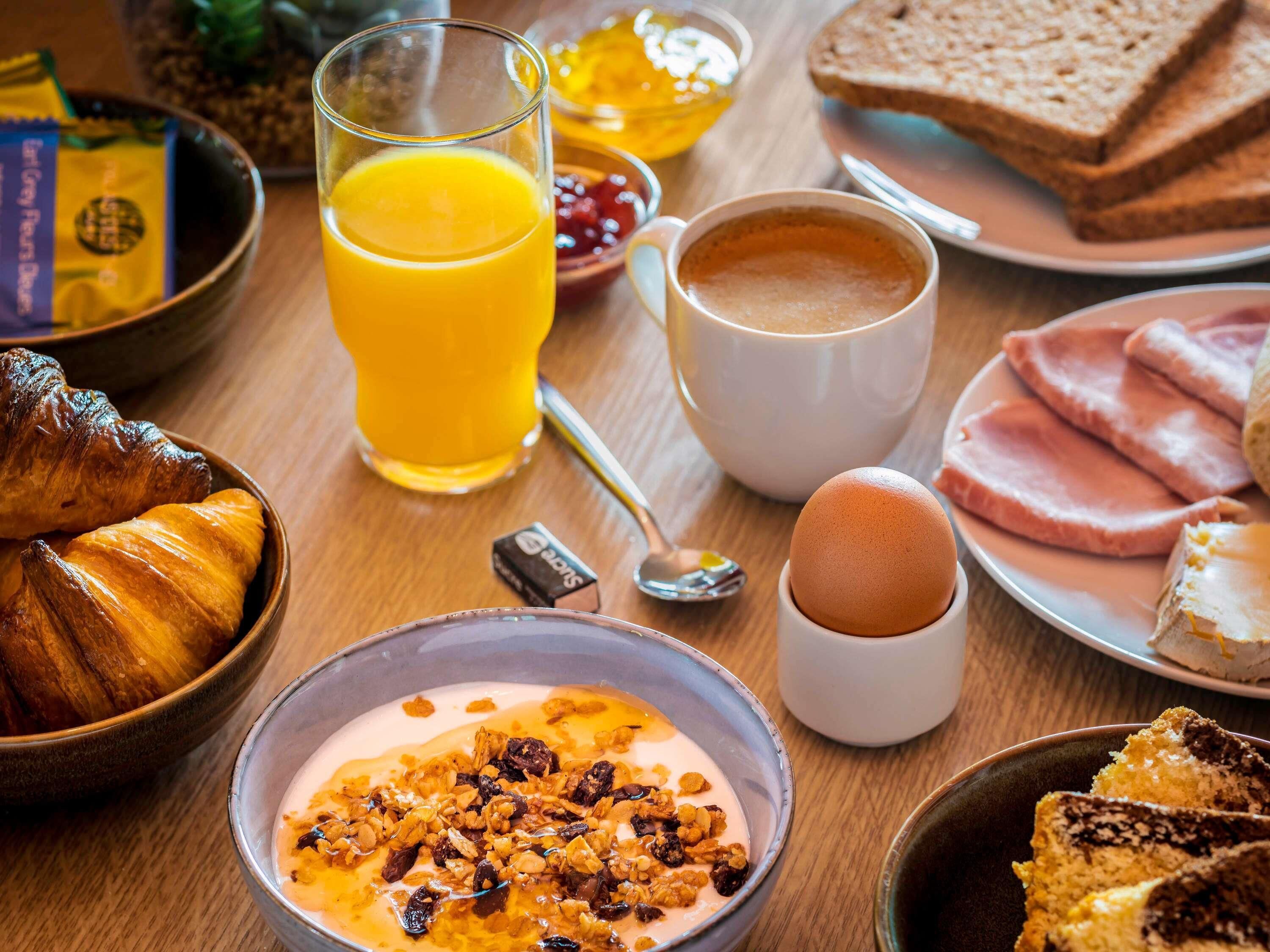 Una mesa de desayuno con cruasanes, jugo de naranja, café, huevos, jamón y tostadas.