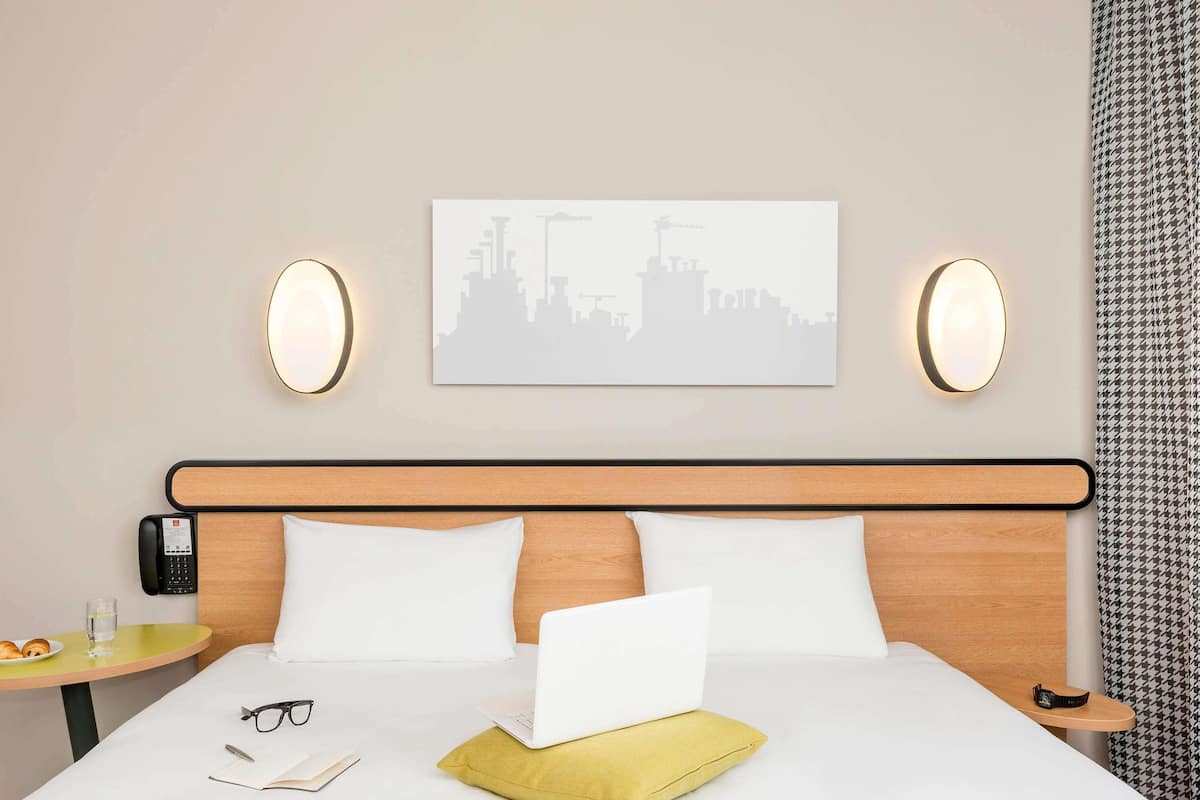 Habitació d'hotel amb un llit, una taula de nit amb un telèfon, un ordinador portàtil, una parella de ulleres i un plat de croissants.