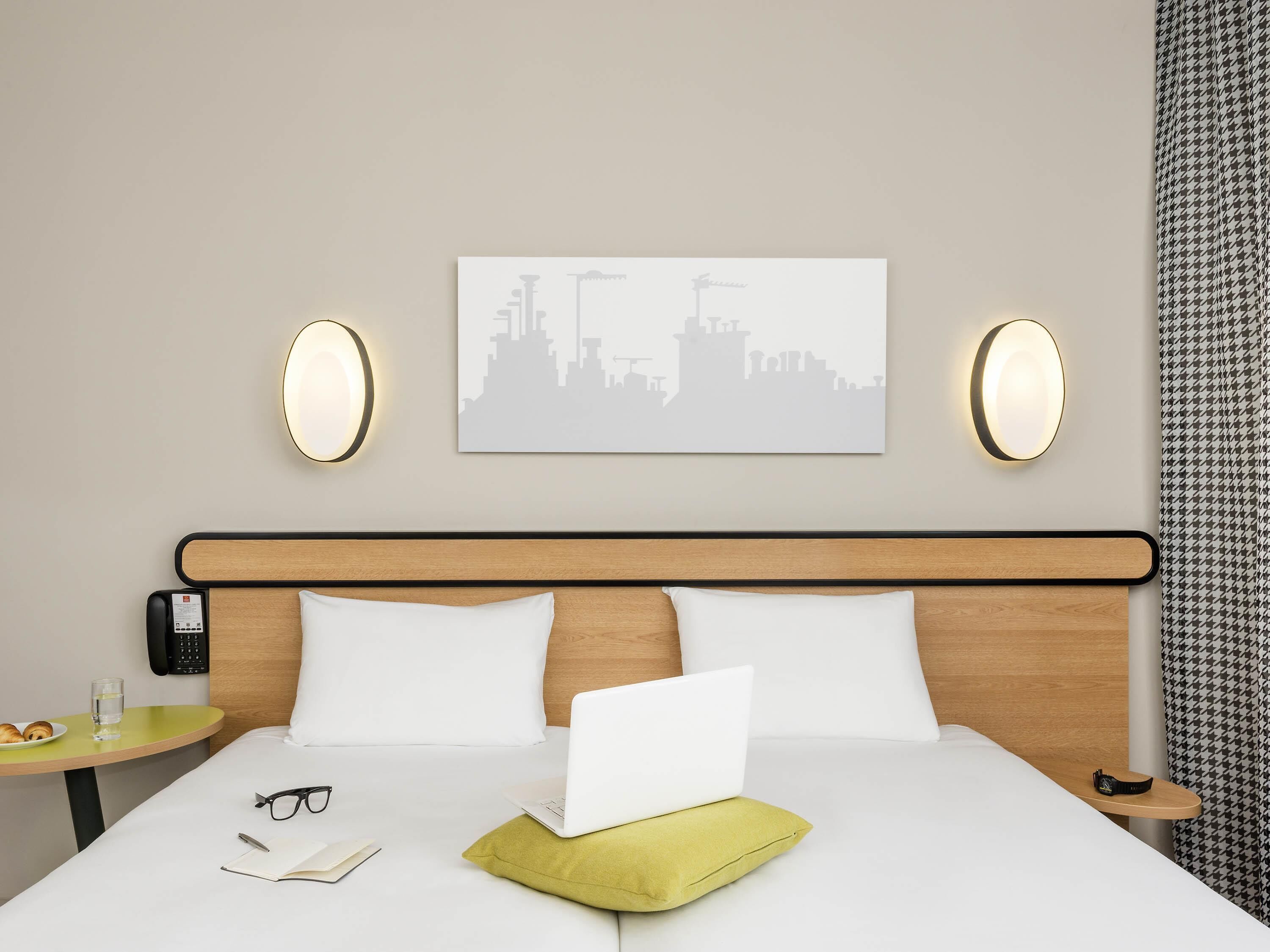 Ein Hotelzimmer mit Bett, Laptop, Notizbuch, Brille, einem Teller mit Croissants, einem Glas Wasser und einem Stadtbild an der Wand.