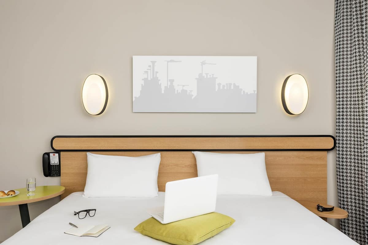 Ein Hotelzimmer mit Bett, Laptop, Notizbuch, Brille, einem Teller mit Croissants, einem Glas Wasser und einem Stadtbild an der Wand.