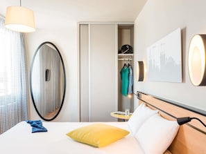 Habitación de hotel con cama, mesita de noche con vaso, perchero y una obra de arte con vista a la ciudad en la pared.