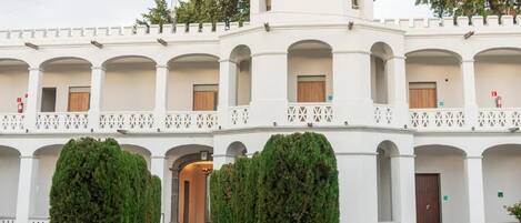 Un patio con un sendero central bordeado por árboles simétricos, que conduce a un edificio blanco con varios balcones y una torre central.