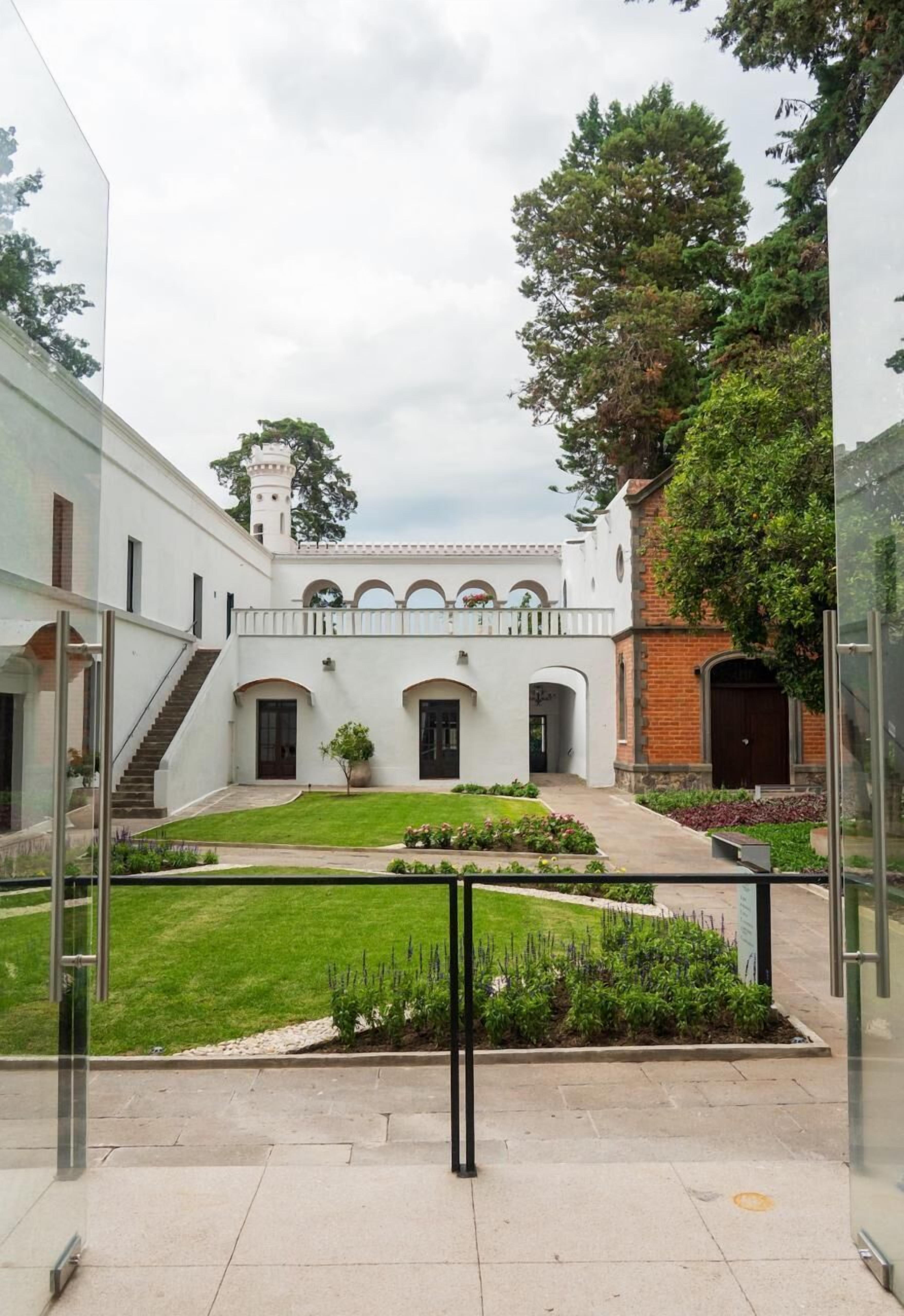 Un patio con jardín central, un edificio blanco con arcos y una construcción de ladrillo con torre.
