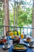Una mesa dispuesta con alimentos para el desayuno, como tortitas, ensalada de frutas y café, con vistas a un área boscosa.