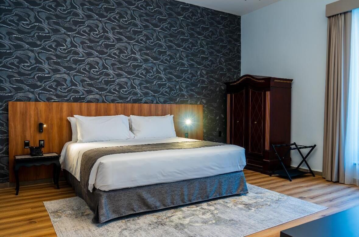 A neatly made bed with white linens, a wooden headboard, and a patterned wall behind it.