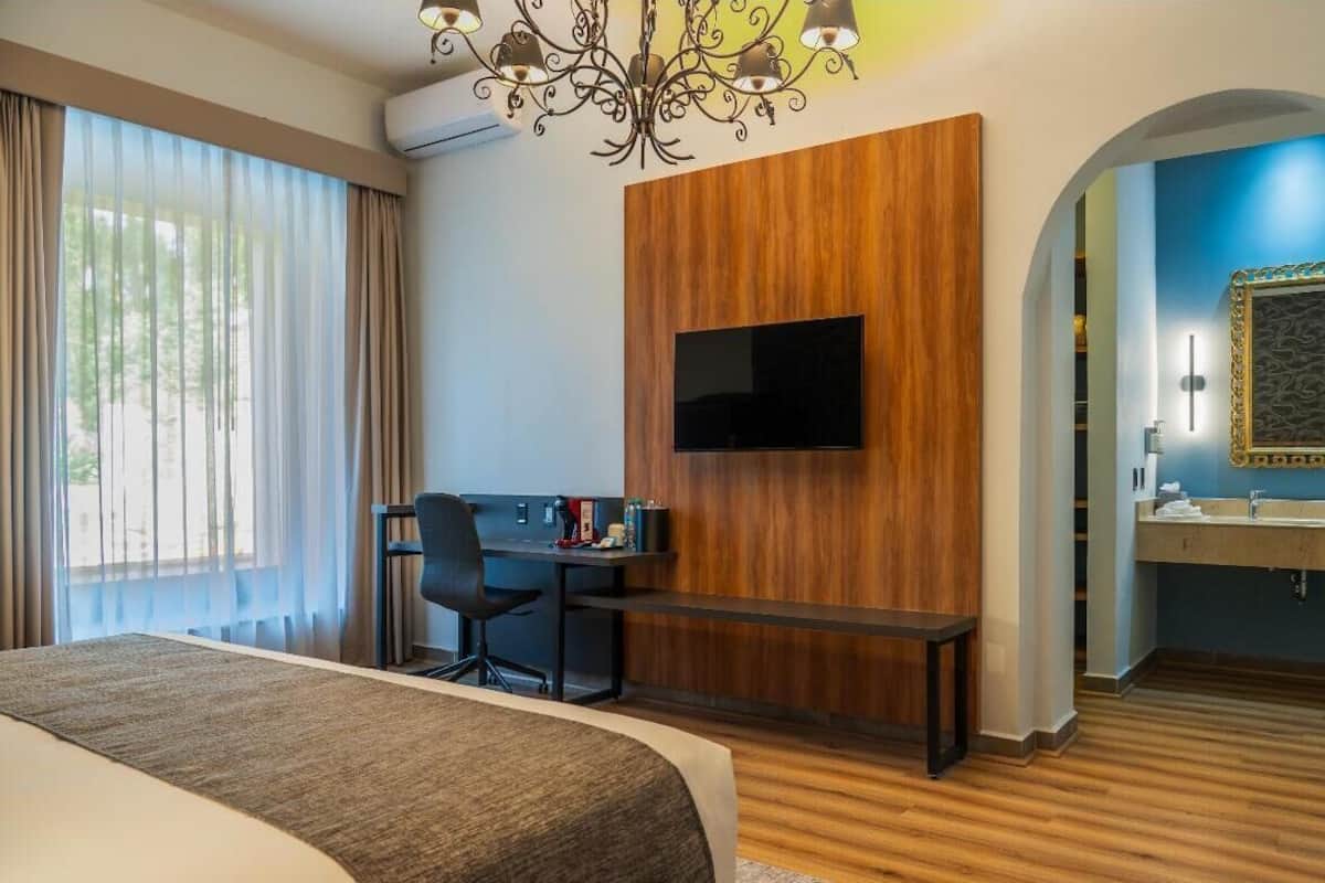 Ein Hotelzimmer mit einem Bett, einem Schreibtisch mit Stuhl, einem an einer Holzplatte montierten Fernseher sowie einem Badezimmer mit Spiegel und Waschbecken.