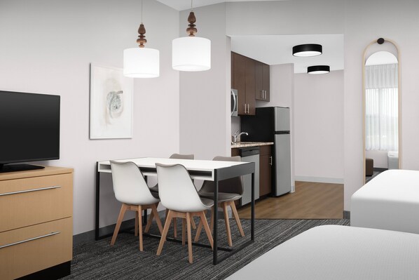 Una habitación de hotel moderna con una televisión de pantalla plana, una zona de comedor con una mesa y sillas blancas, una cocina con armarios y un refrigerador, y un espejo grande.
