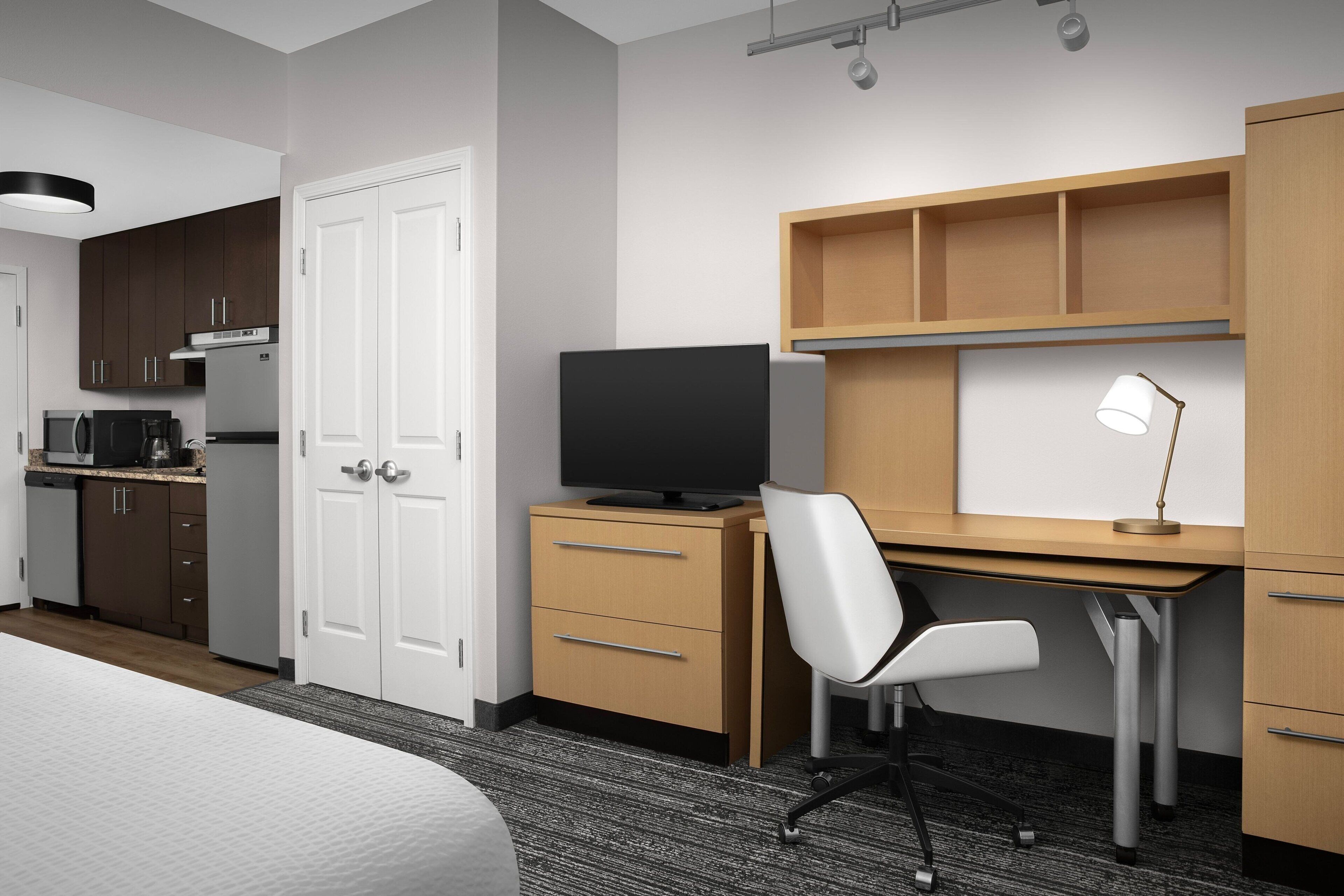 A hotel room with a bed, a desk with a computer and lamp, a TV, a kitchenette, and a wardrobe.
