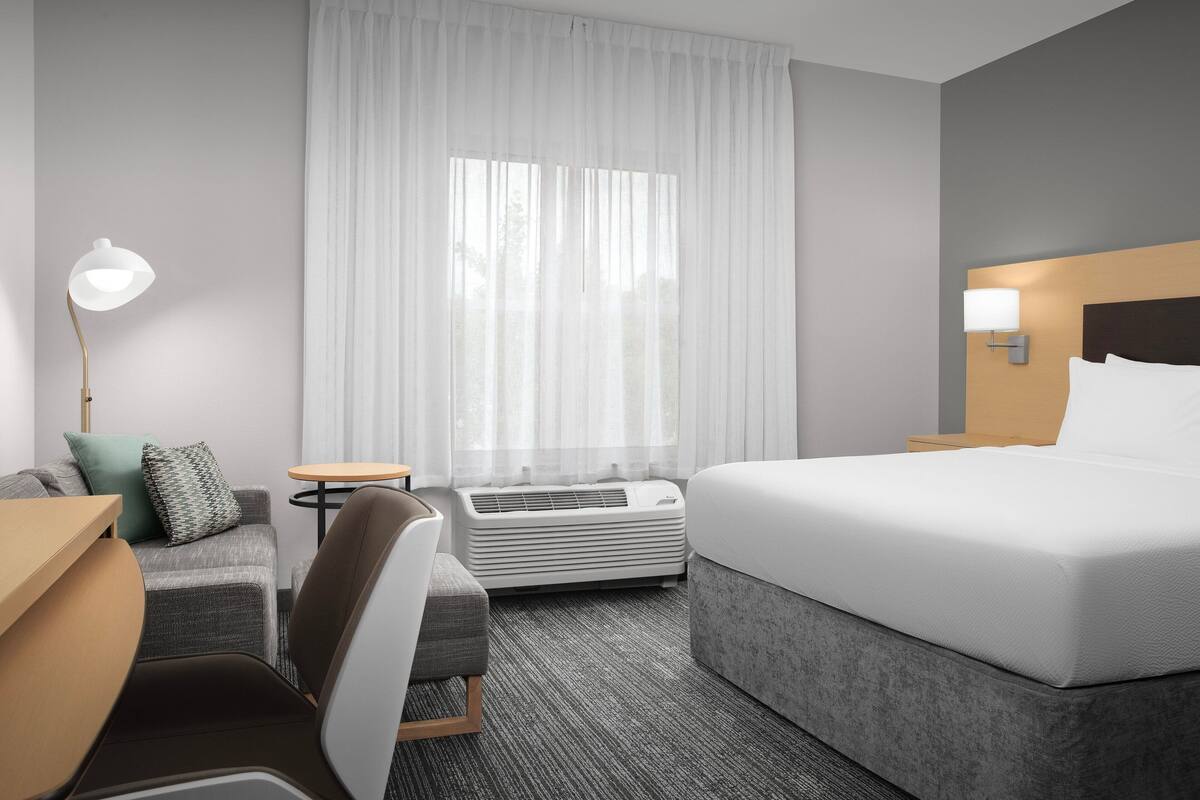 A hotel room with a bed, a desk, a chair, a lamp, a small table, and a window with curtains.