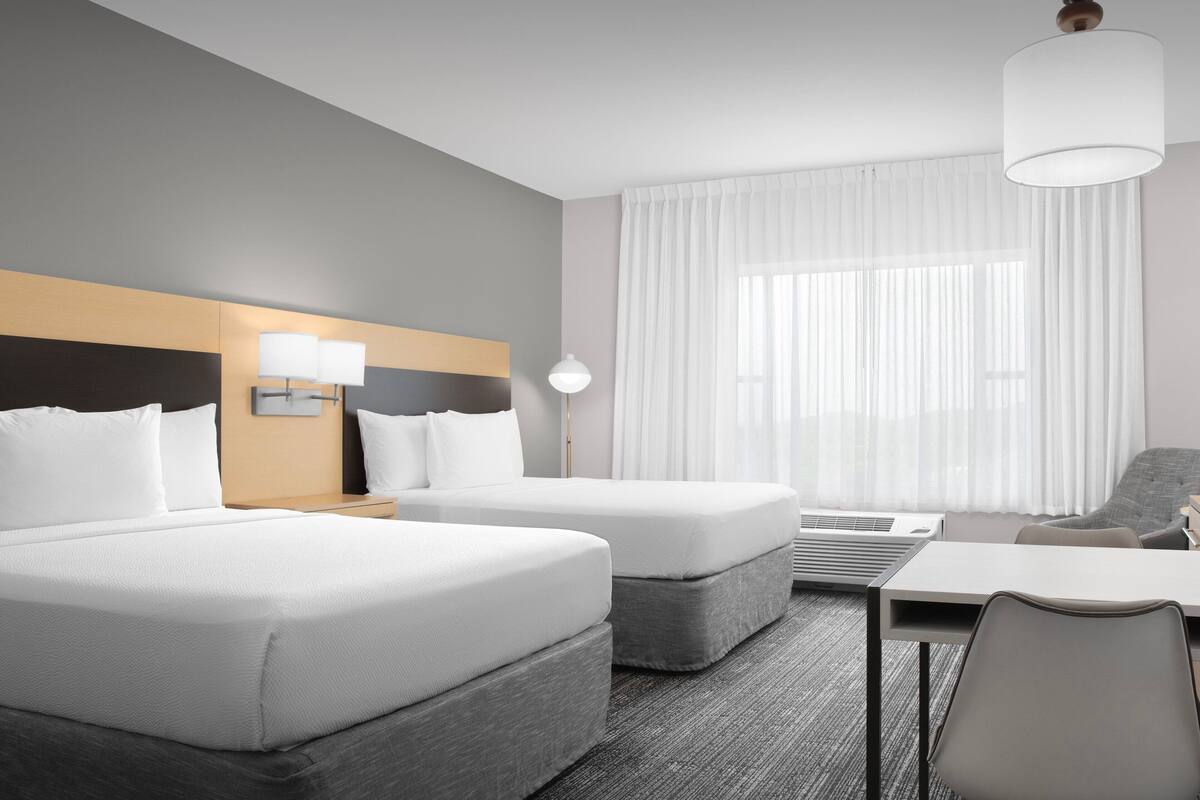 A hotel room with two beds, a desk, a chair, a lamp, and a large window with curtains.