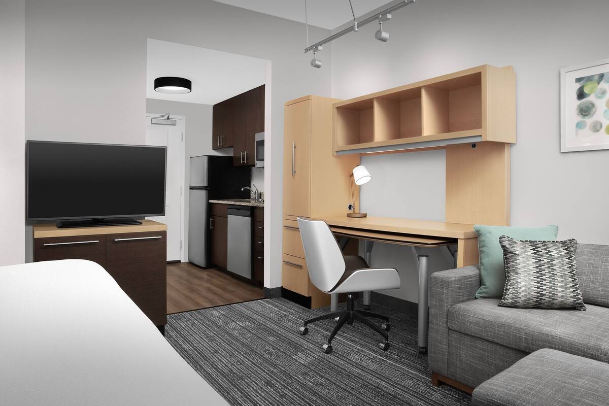 A modern hotel room with a flat-screen TV, a desk with a lamp, a grey sofa with a patterned pillow, and a kitchenette.