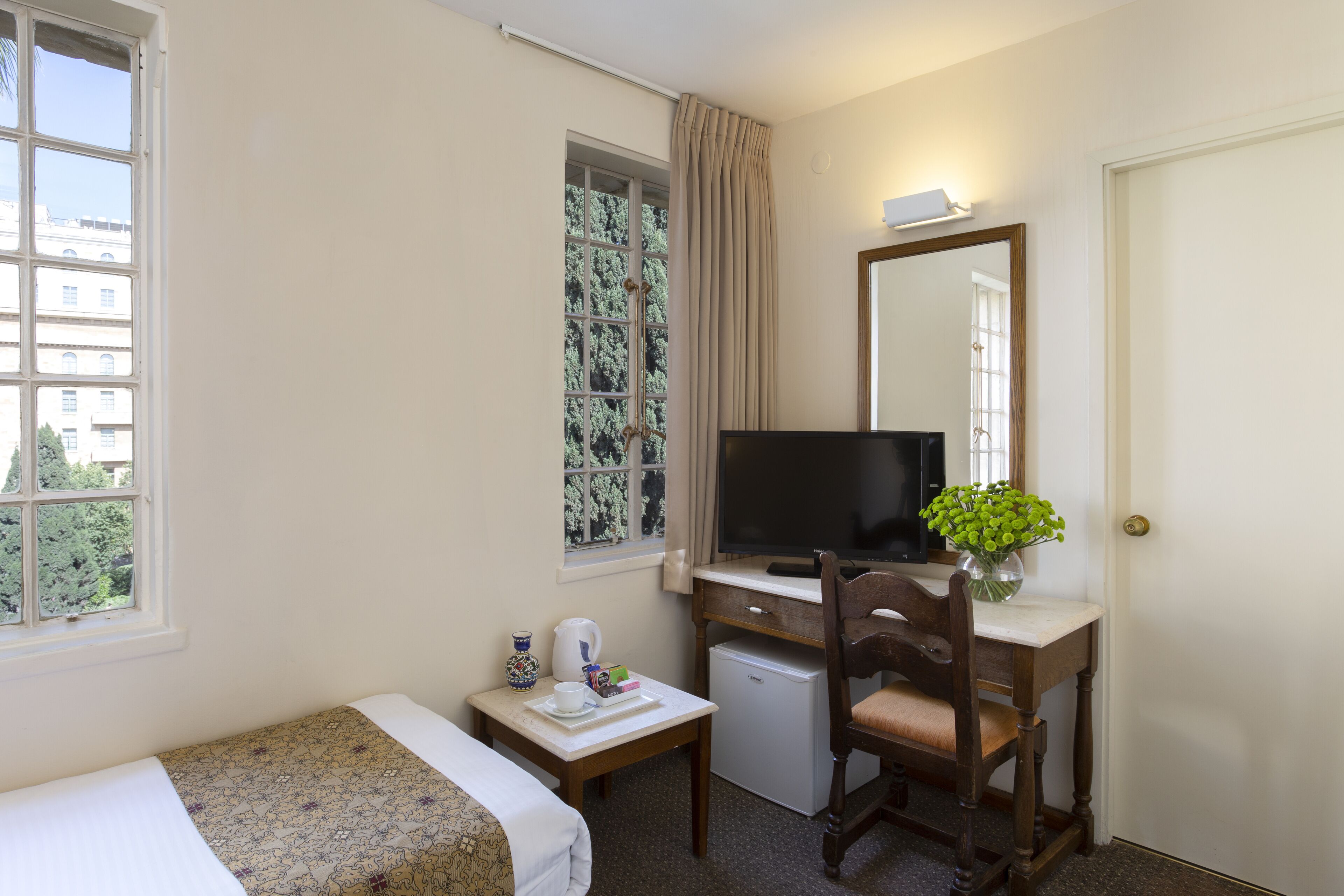 Een hotelkamer met een bed, een bureau met een computer, een kleine koelkast, een stoel, een raam met gordijnen en een deur.