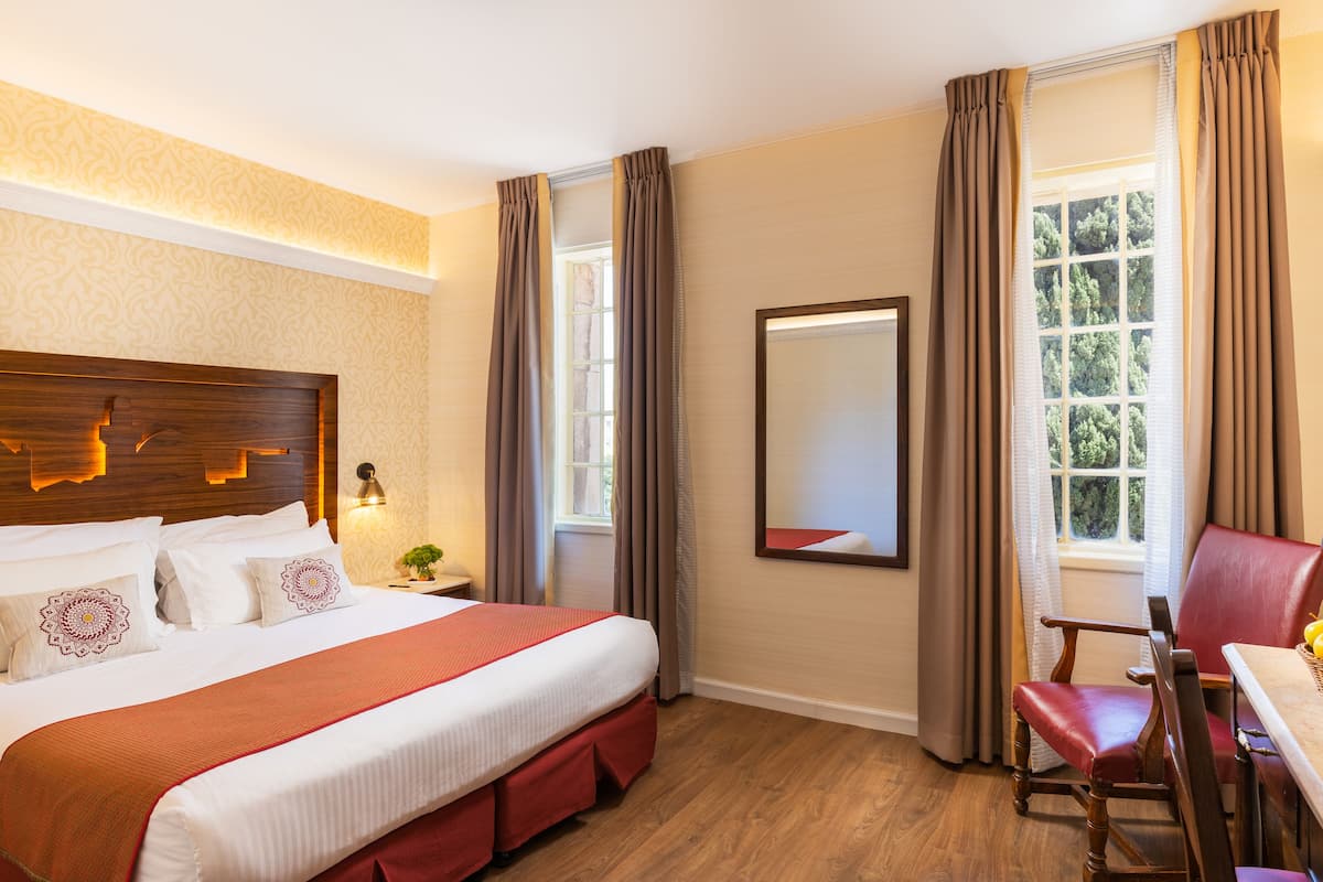 Une chambre d’hôtel avec un grand lit, une tête de lit en bois, un bureau avec une chaise, un miroir et une fenêtre avec des rideaux.