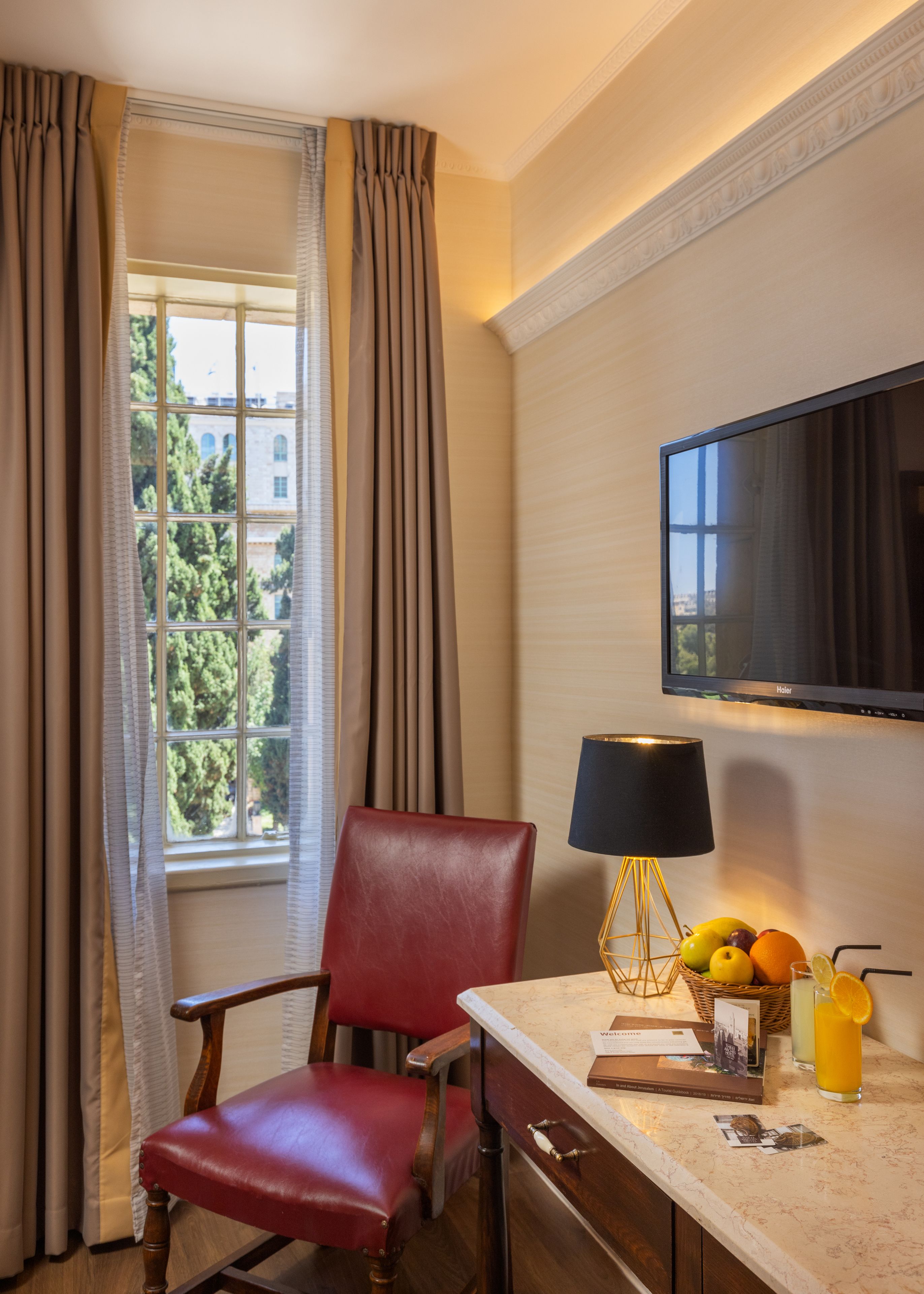 Habitación de hotel con una butaca de cuero rojo, un televisor de pantalla plana en la pared, una mesa con una lámpara, una cesta de frutas y un vaso de jugo de naranja.