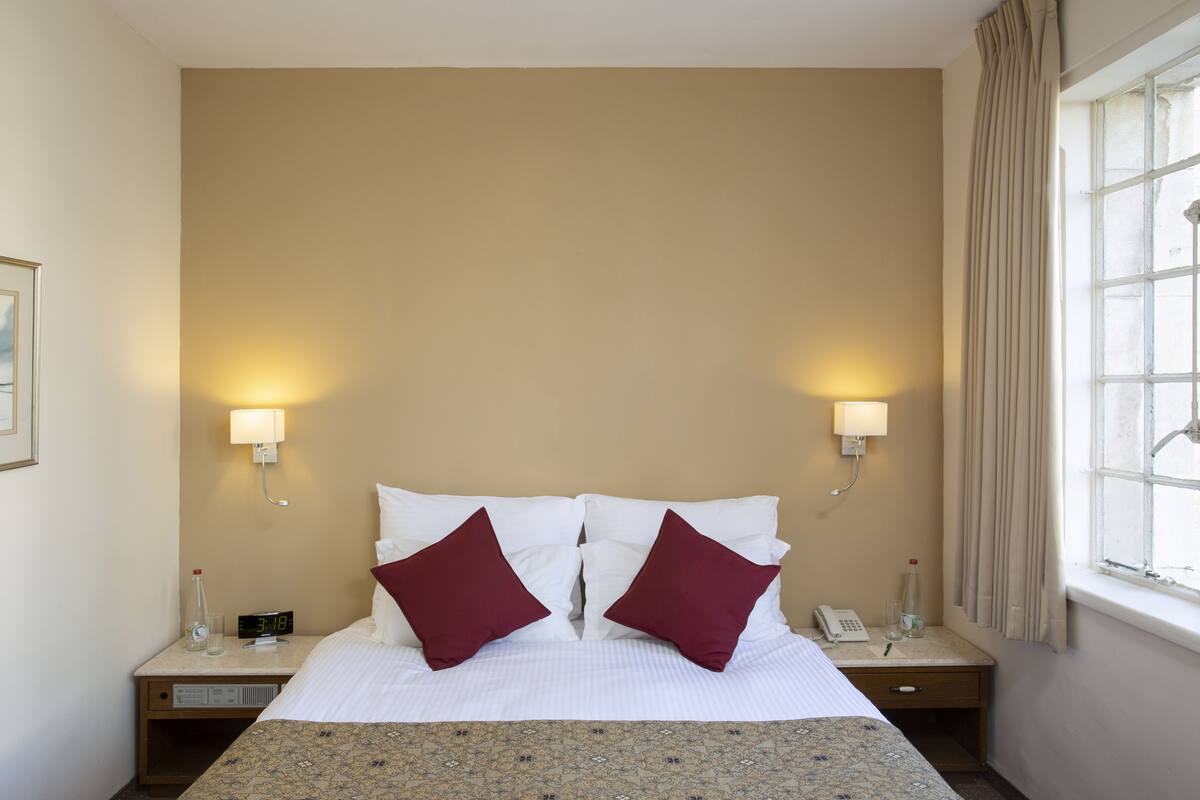 Une chambre d’hôtel comprenant un lit, deux lampes de chevet, une table de chevet, une horloge numérique, un téléphone et une fenêtre avec des rideaux.