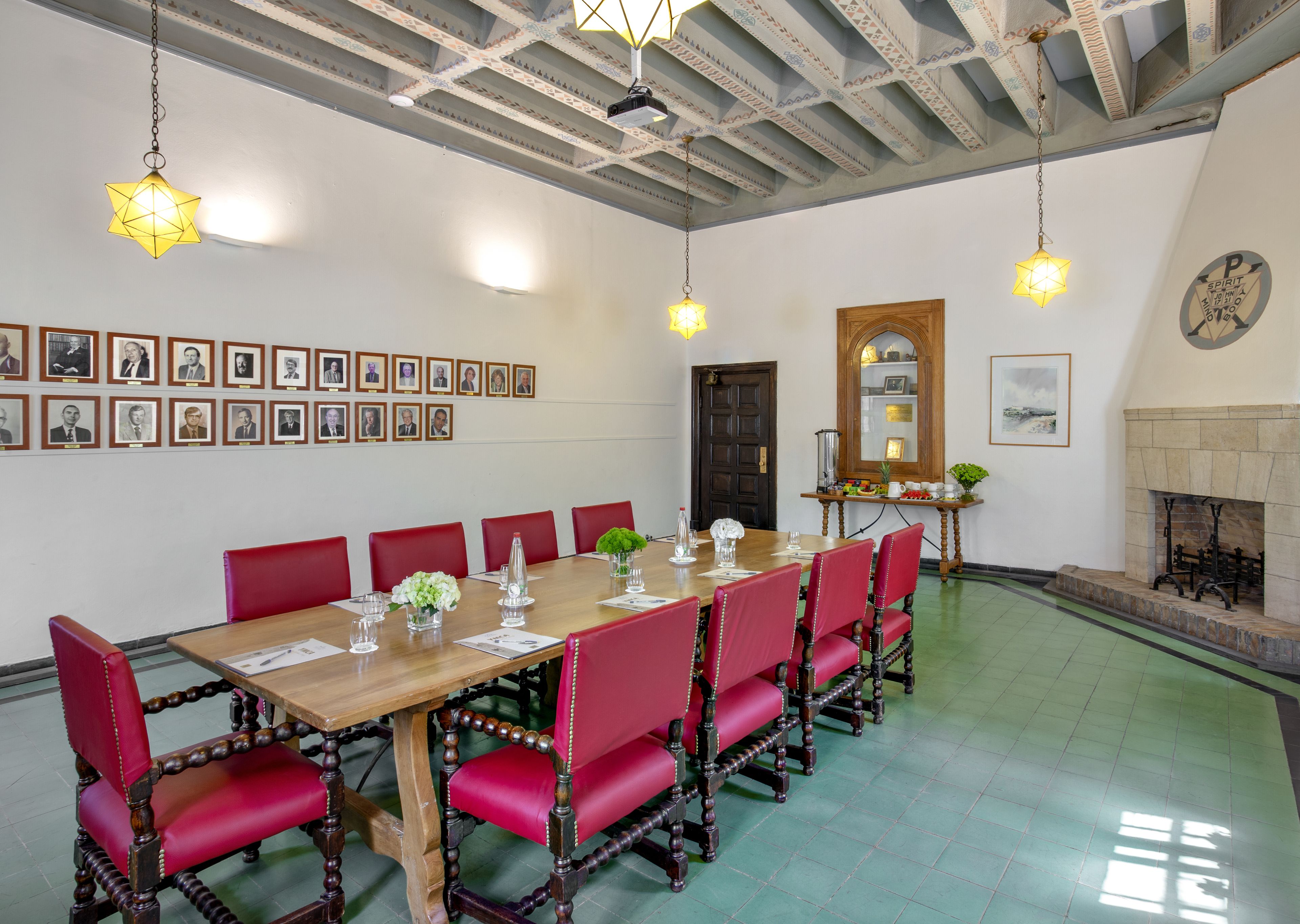 A conference room with a long wooden table, red upholstered chairs, framed portraits on the wall, and a fireplace.