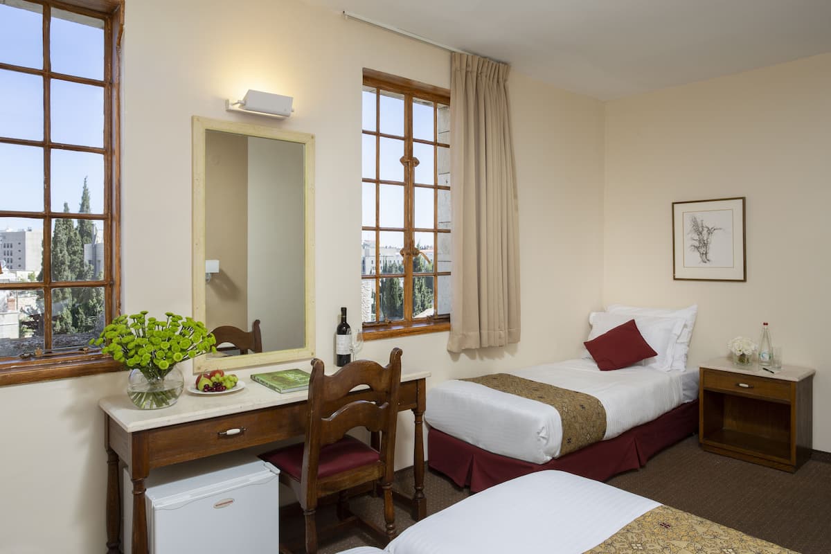 Une chambre d’hôtel avec deux lits, un bureau avec une chaise, un petit réfrigérateur, un miroir et une fenêtre donnant sur des immeubles.