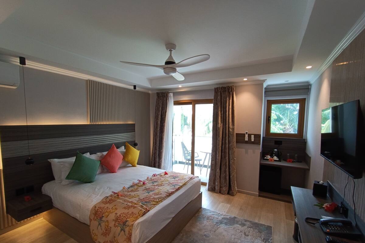 Une chambre d’hôtel moderne dotée d’un grand lit, d’un ventilateur de plafond, d’une télévision et d’un balcon offrant une vue sur la verdure.