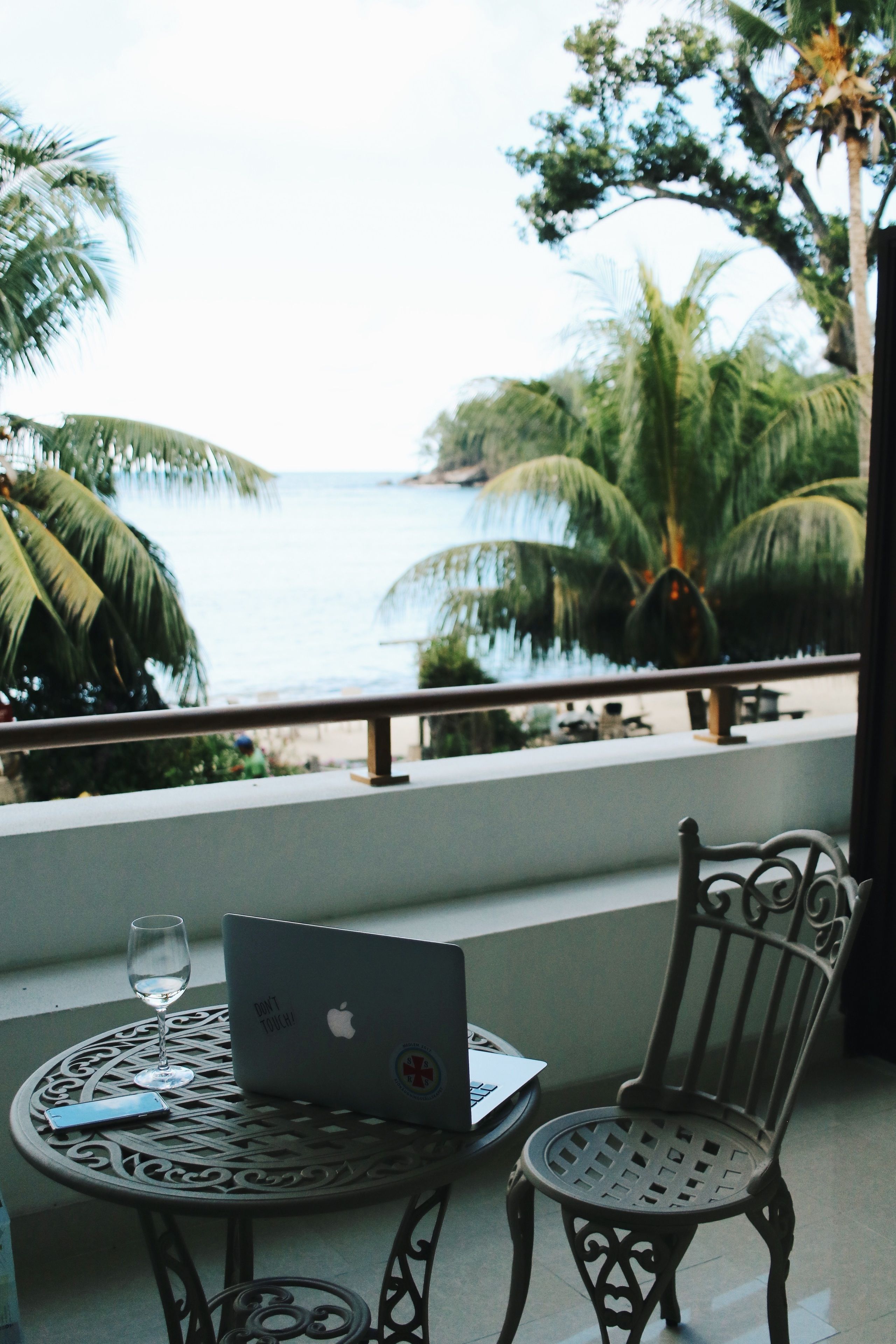 A balcony with a laptop, a glass of water, and a chair overlooking a beach with palm trees.