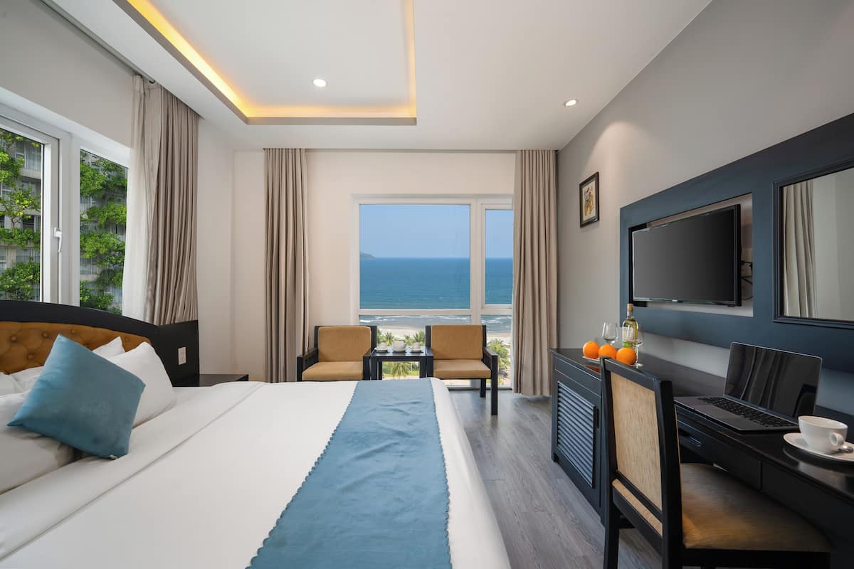 A hotel room with a bed, two chairs, a desk with a laptop, a TV, and a view of the sea.