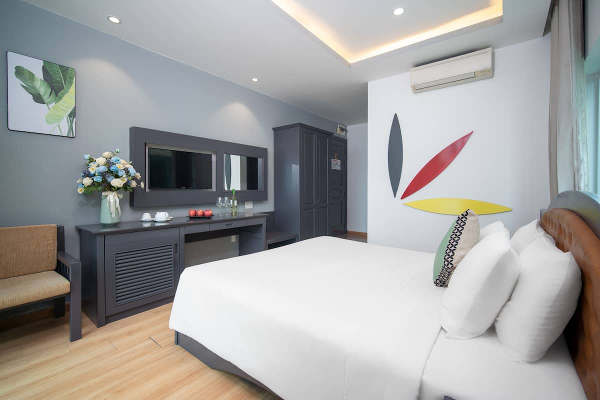 A modern hotel room with a large bed, a desk, a chair, a television, and a wall-mounted air conditioner.