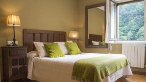 Una cama bien hecha con colcha blanca y verde, una mesita de noche de madera con lámpara, y un espejo grande que refleja una vista de bosque.