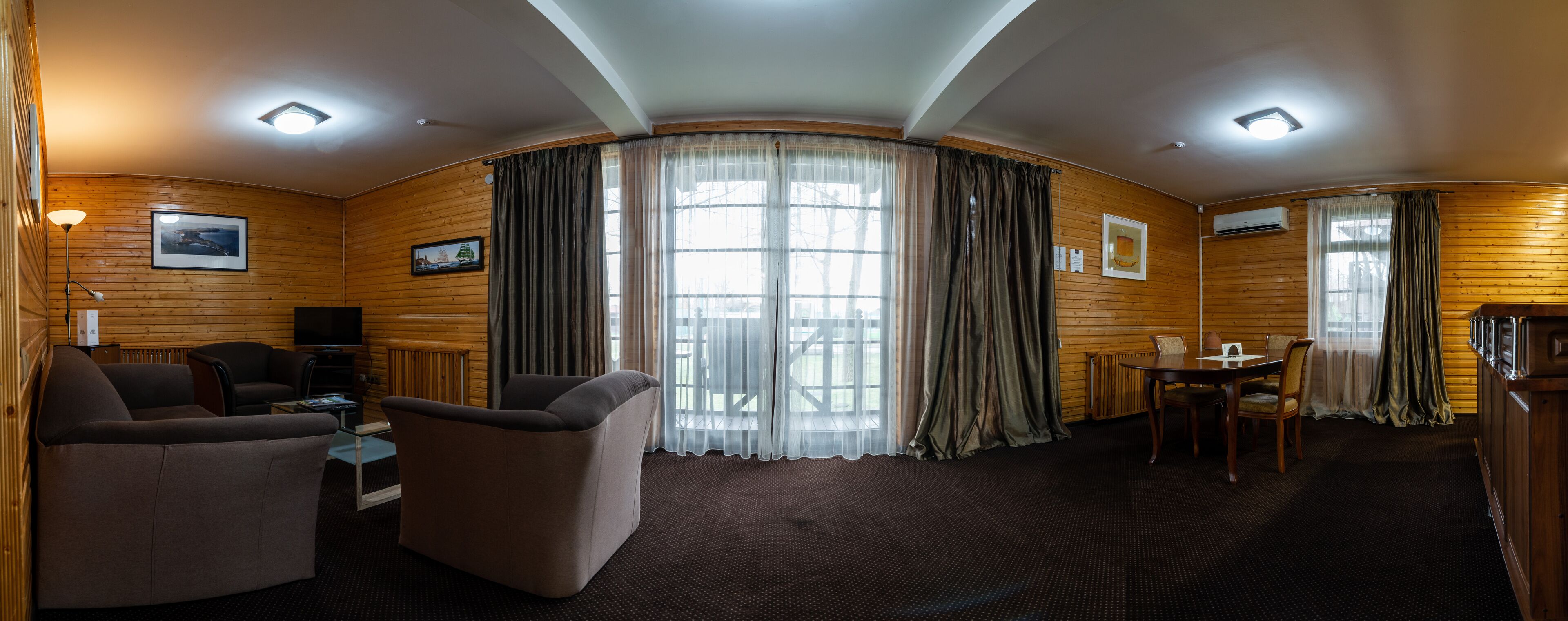 Habitación de hotel con interior de madera, un sofá, un televisor, una mesa de comedor con sillas y ventanas grandes con cortinas.