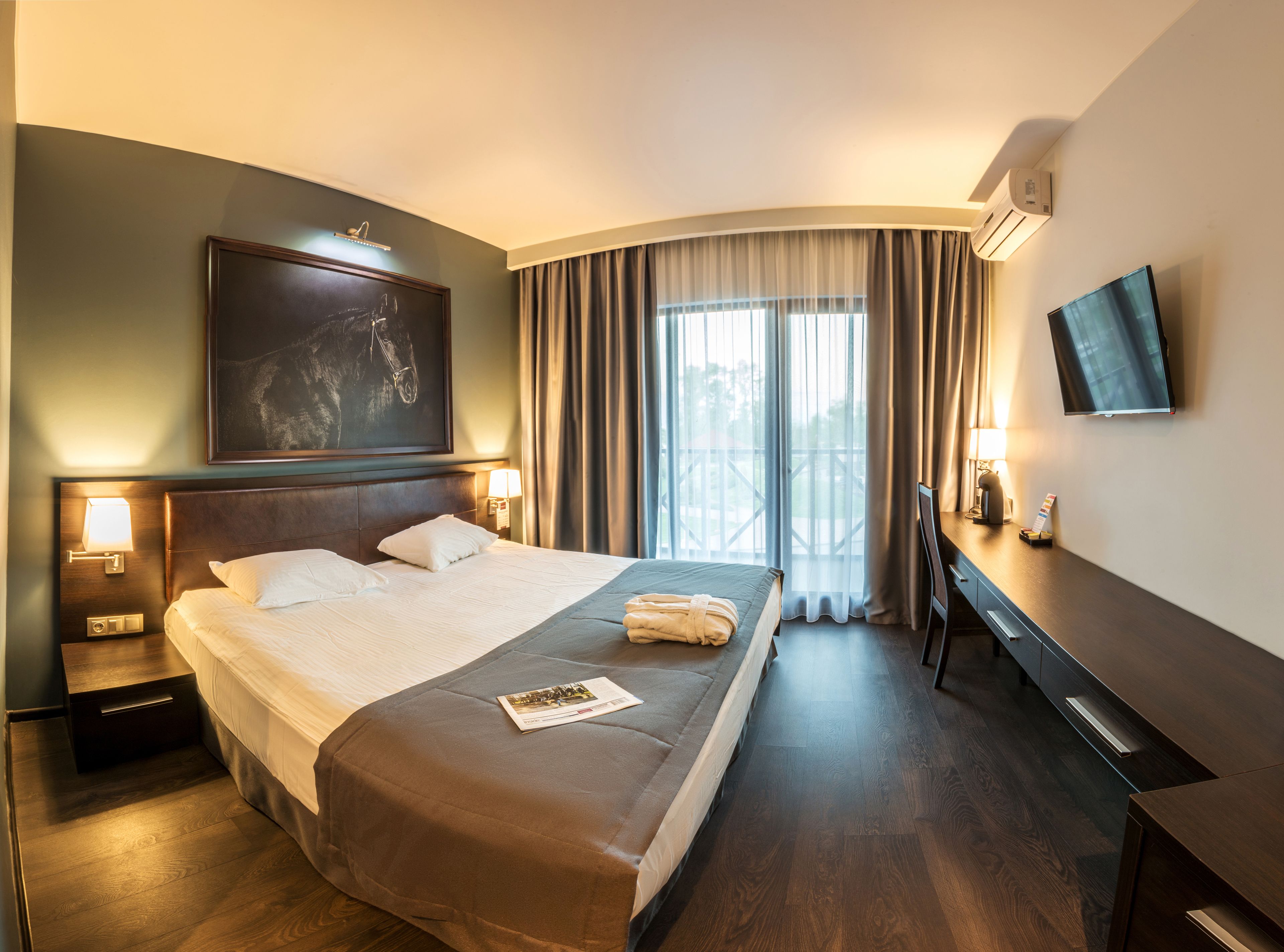 A hotel room with a large bed, a desk, a flat-screen TV, and a balcony with curtains.