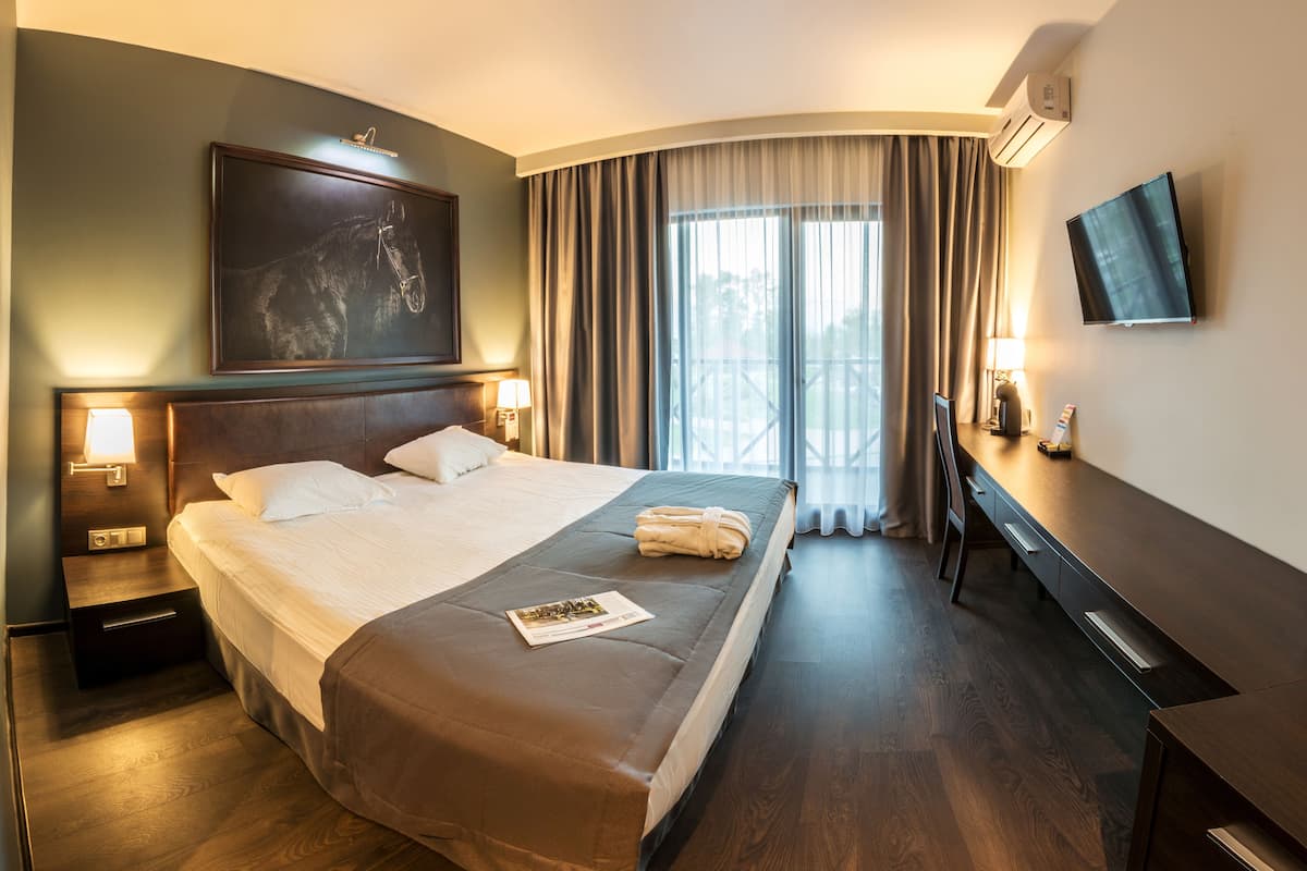 Ett hotellrum med en stor säng, ett skrivbord, en platt-TV och en balkong med gardiner.