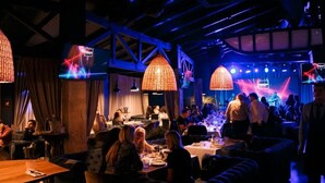 A dimly lit restaurant with tables and chairs, large hanging lights, and a stage with performers.
