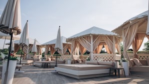 A covered outdoor seating area with cushioned benches and umbrellas, surrounded by wooden lattice walls and potted plants.