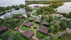 An aerial view of a lakeside resort with multiple wooden cabins, a marina with boats, and a sports field.