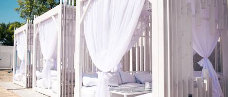 Una hilera de cabañas de playa blancas y cerradas, con asientos acolchados y una mesa de vidrio, cada una con un dosel.