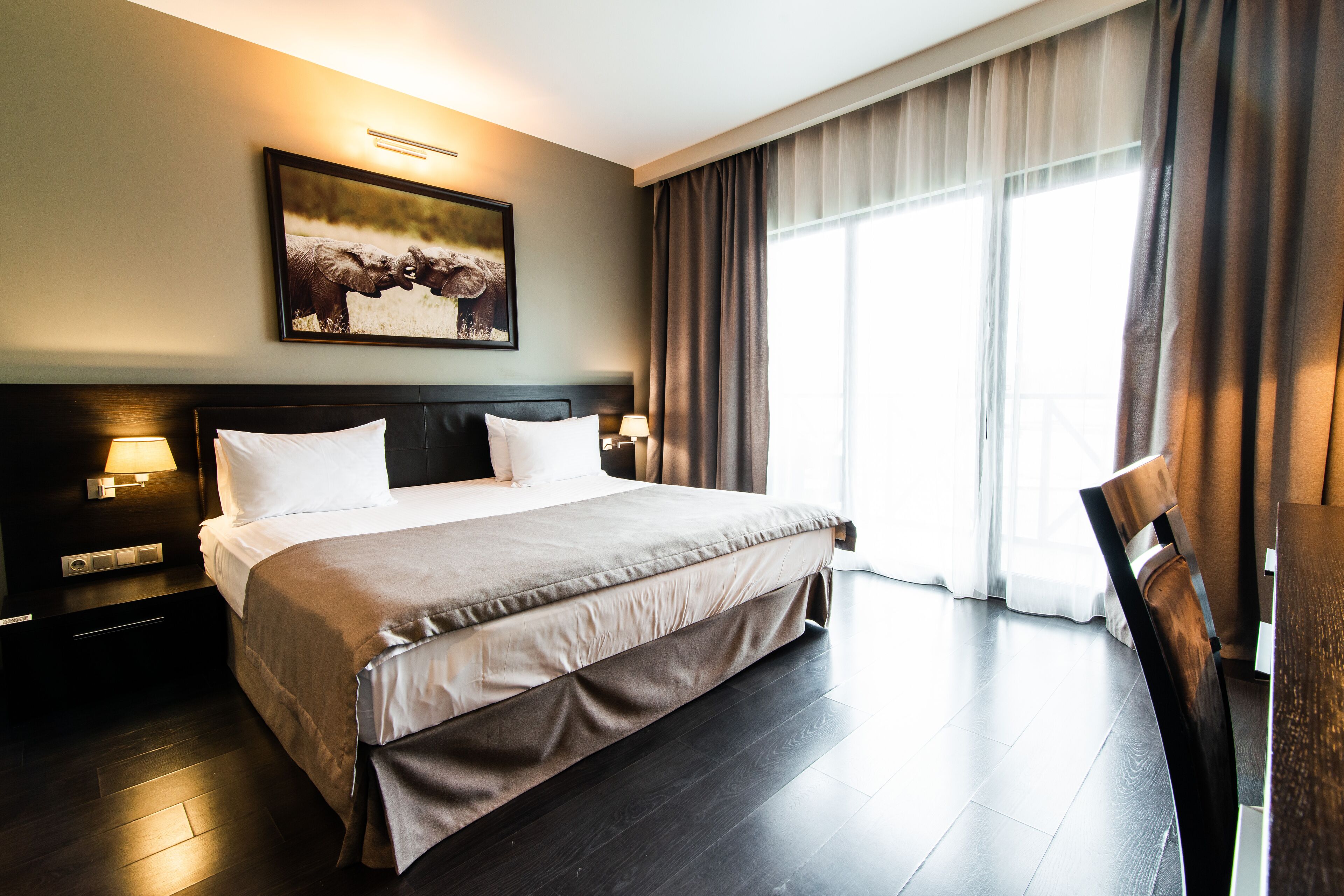 Habitación de hotel con una cama grande, una mesita de noche con lámpara, un cuadro en la pared y una ventana con cortinas.