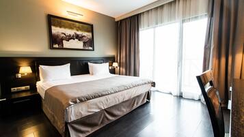 Habitación de hotel con una cama grande, una mesita de noche con lámpara, un cuadro en la pared y una ventana con cortinas.