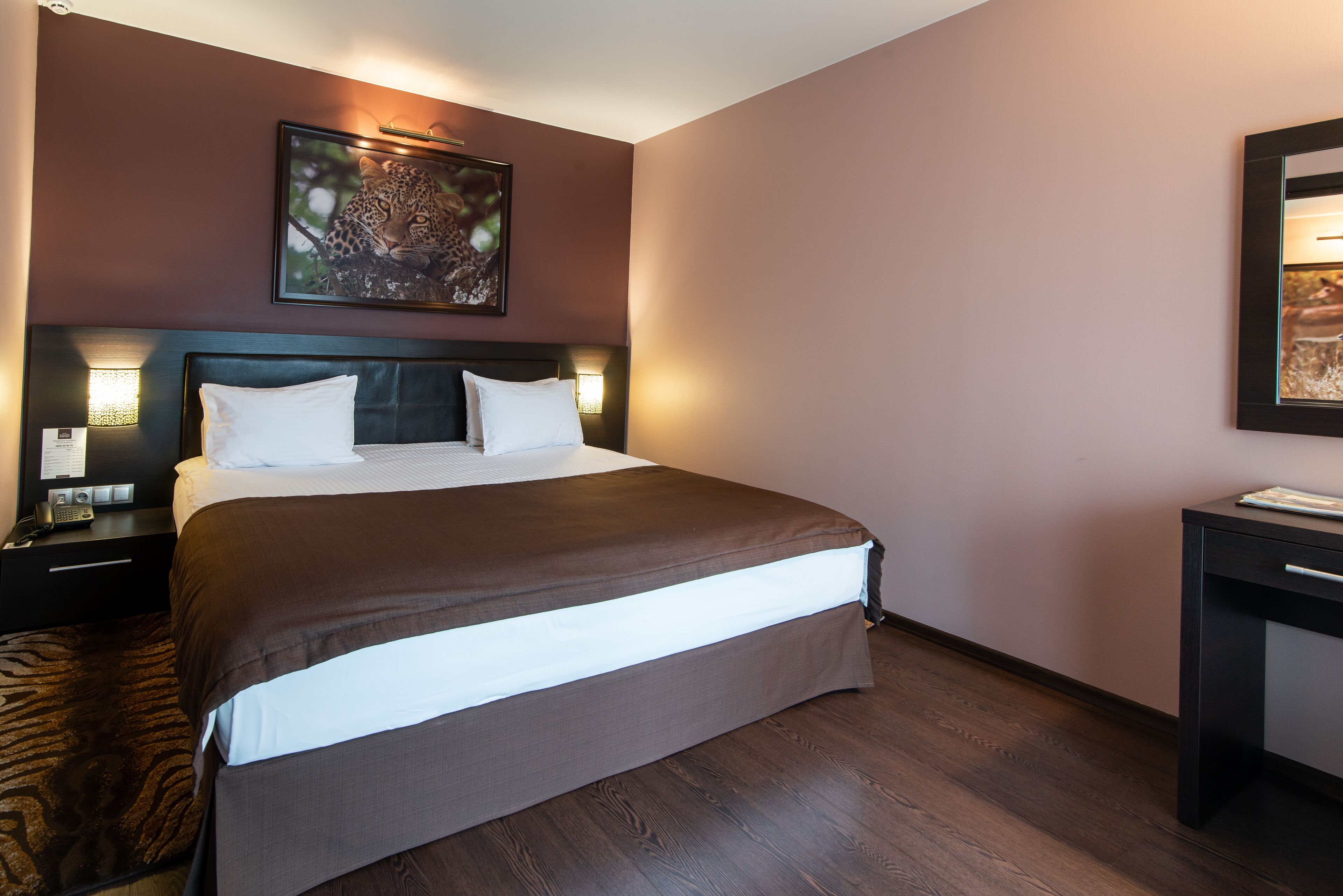 A hotel room with a large bed, dark wood flooring, a headboard with a light fixture, a nightstand with a phone, and a desk with a mirror.