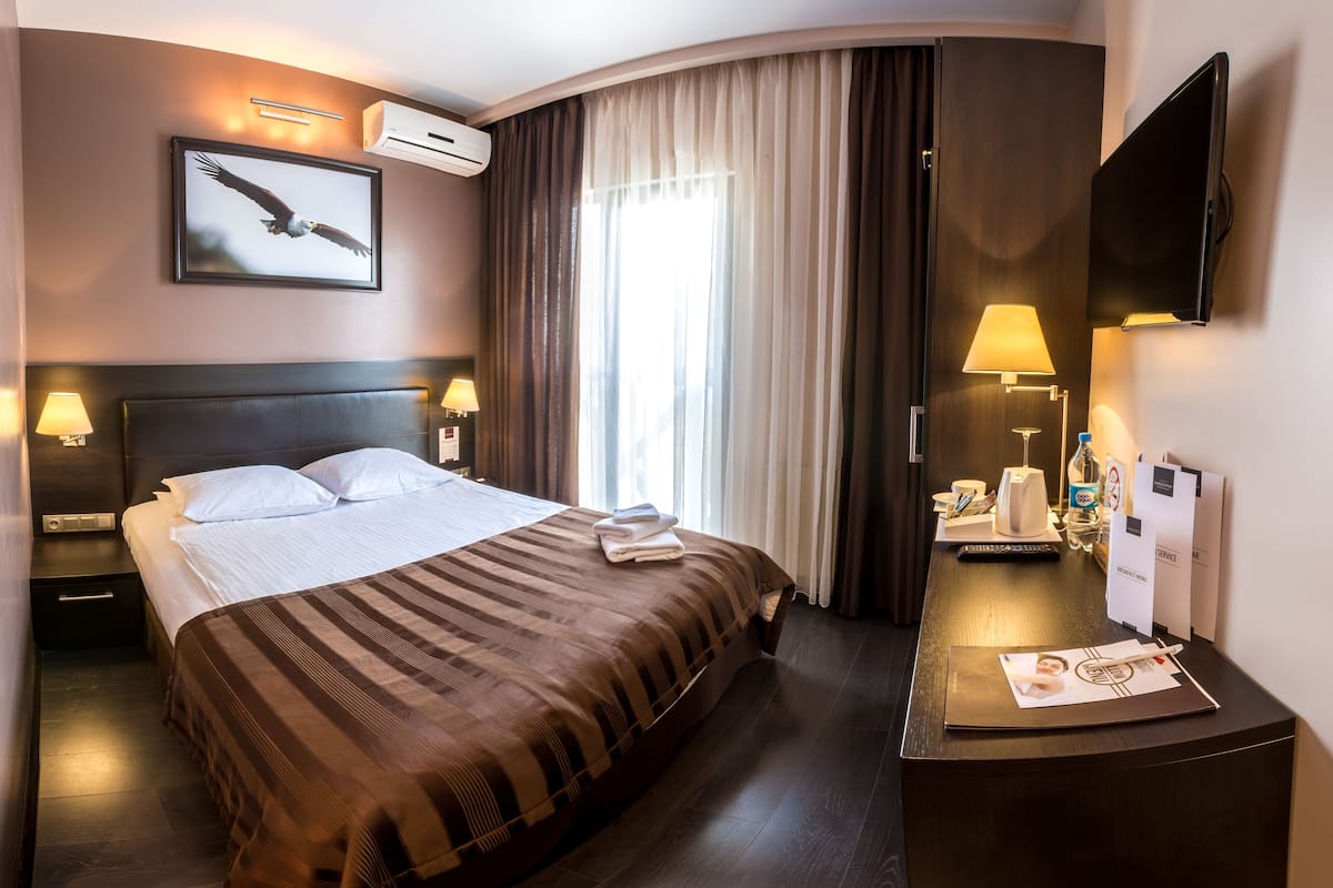 Ett hotellrum med en stor säng, en platt-TV, ett skrivbord med en lampa, en vattenflaska och en inramad tavla på väggen.