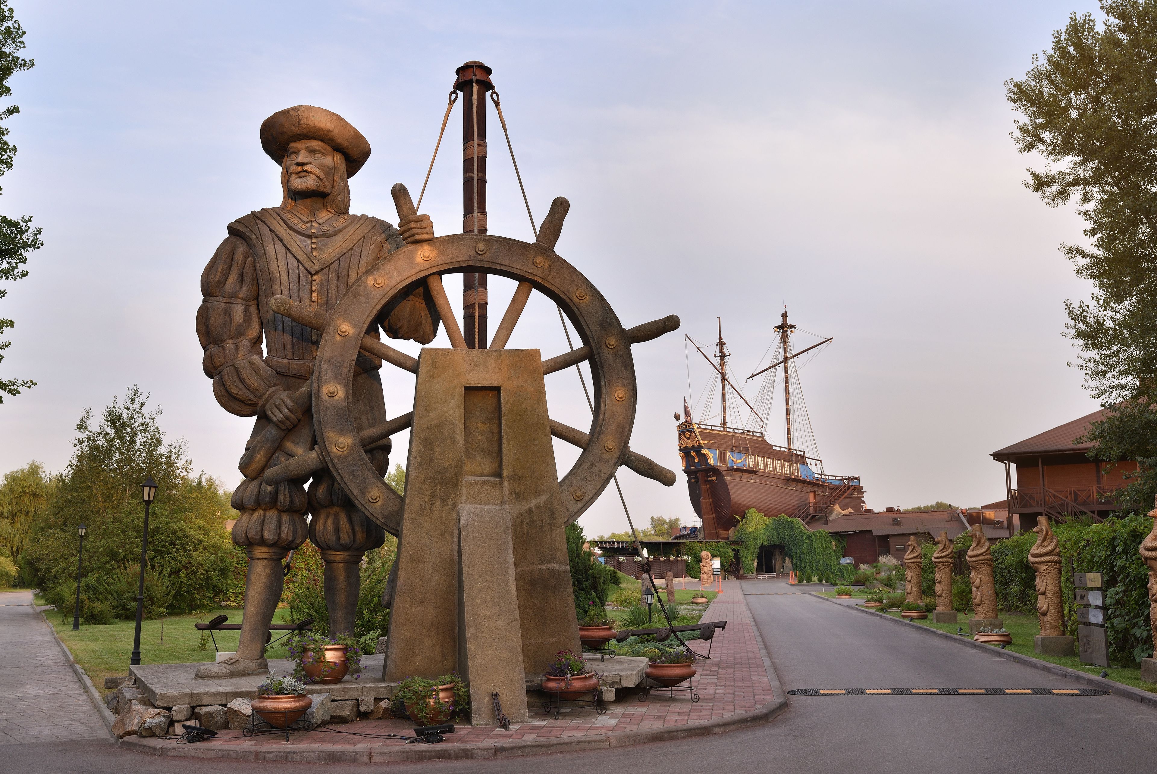 A large outdoor statue of a historical figure with a ship's wheel, surrounded by potted plants and a pathway.